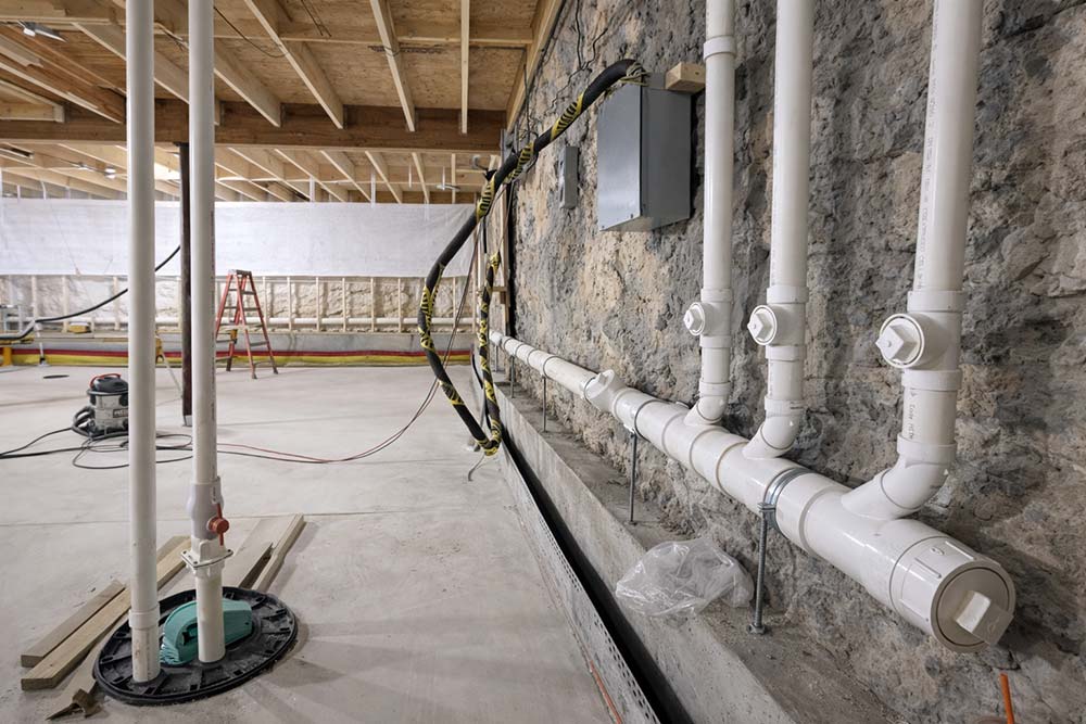 New construction basement plumbing installation with white PVC drain piping secured along stone foundation wall.
