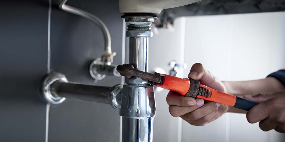 Chase Plumbing Co. - Local Plumbing Expert Plumber tightening a sink drain pipe connection with a wrench under a bathroom sink.